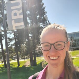 A woman with blonde hair wearing glasses and a pink short smiles in the foreground; Thompson Rivers University campus is in the background.