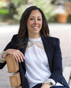 Image of a woman with brown hair, a white shirt, and navy jacket seated on a bench